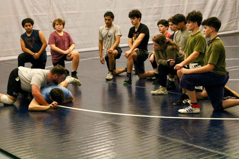 Sussex Academy grapplers (l-r) Mizeal Guevara Yoc, Asher Kern, Elean Roblero, Will Hendershot, Ryan Peña, Talah Patton, Jaxon Dufour, Cleverson Ortiz, Charlie Bowman and Joel Denham watch head coach Billy Ott as he shows them a move on assistant coach Cory Lawson.